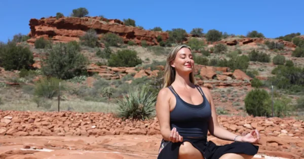 SWR_jolene -hero image A woman meditates cross-legged on red rocks, hands on knees, eyes closed. Green shrubs and Sedona’s red formations under blue sky.