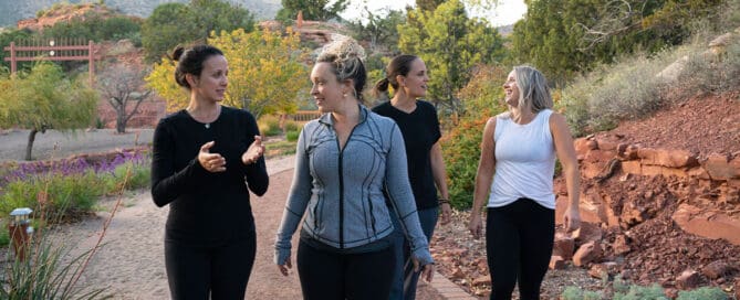 Group of women walking and talking on a scenic desert path at a retreat center, surrounded by red rocks and greenery.