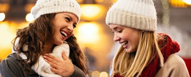 Two women wearing knit hats and scarves laughing joyfully outdoors during the winter holiday season.