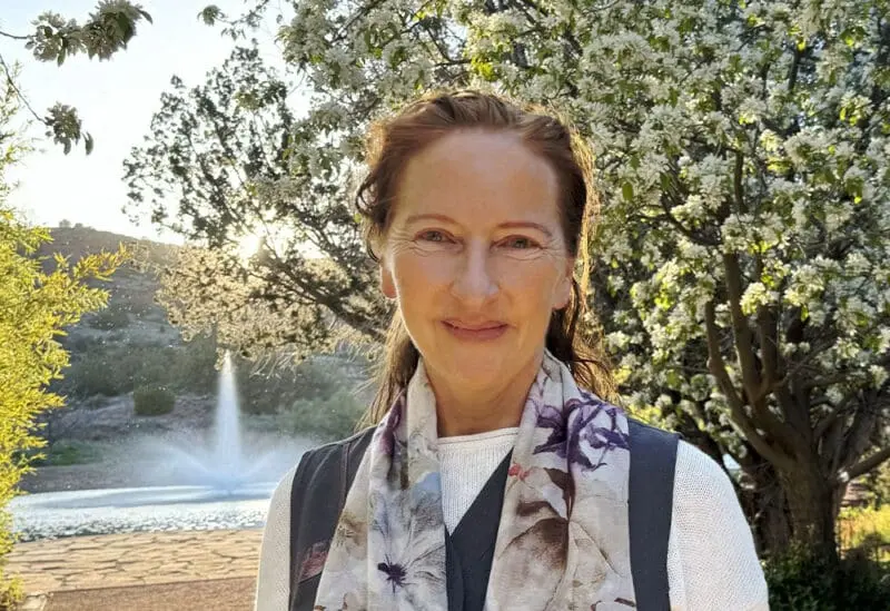 Beneath sunlit blooms, a woman with light brown hair, floral scarf, and white sweater smiles peacefully by a fountain outdoors.