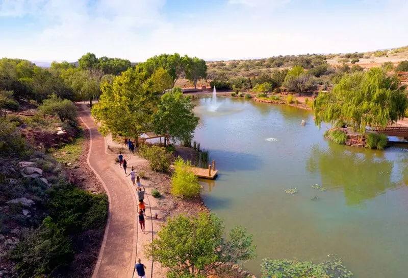 A peaceful path curves beside a shimmering pond and fountain, embraced by lush trees; people stroll under the warm sun’s light.