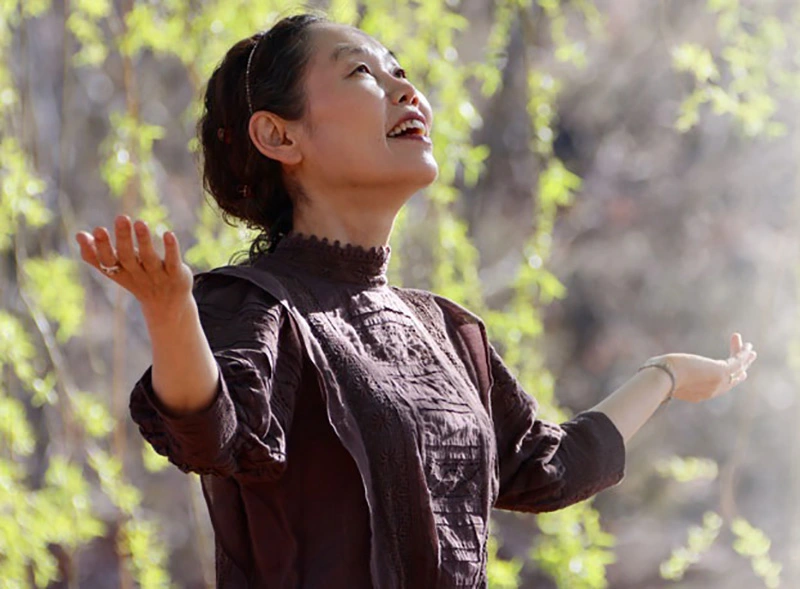 trainer_jahbi-800 A woman in a brown long-sleeve top stands outside, arms raised, smiling up with joy amid sunlit green leaves and uplifting energy.