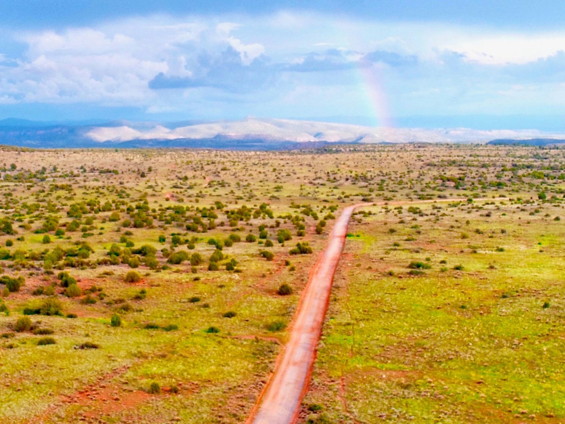 founding-day-bill-gray-cleanup-2 A dirt road winds through a lush green desert beneath clouded skies, a soft rainbow appearing gently by distant rolling hills.