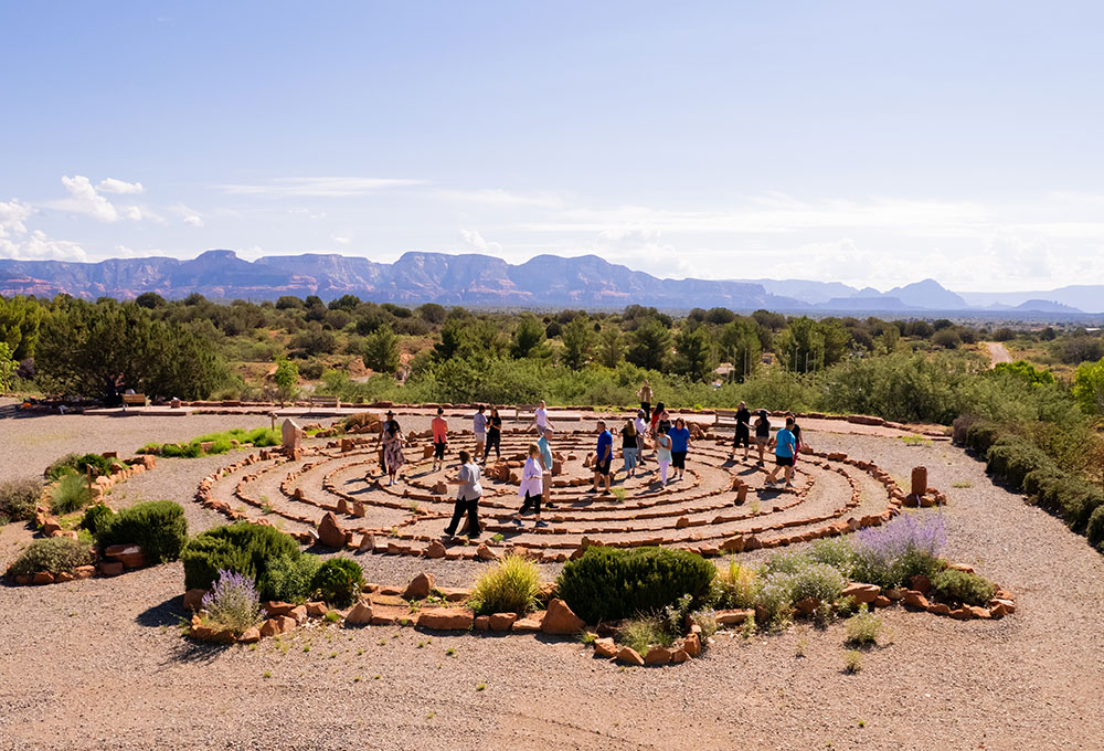 community-donate People walk quietly through a stone labyrinth in the desert, embraced by green shrubs, mountains, and an expansive blue sky.