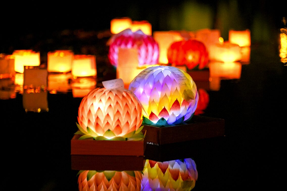 Lanterns of many shapes, aglow in orange, pink, purple, and yellow, softly float on night waters, their light mirrored below.