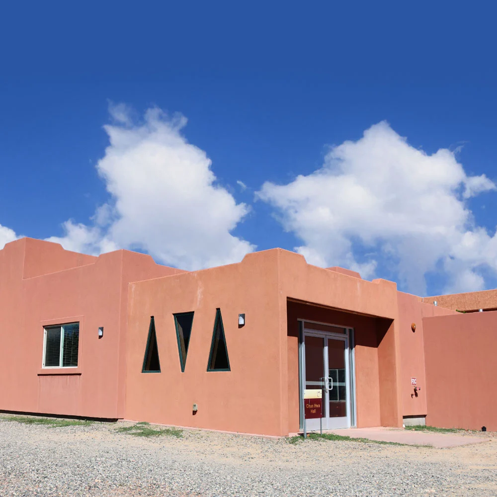 A serene adobe-style building with smooth, pink-brown walls and glass doors sits beneath a vibrant blue sky, gravel stretching before it.