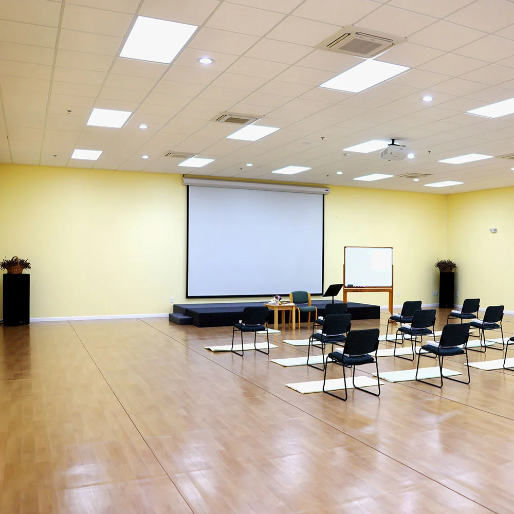 A bright, peaceful room with pale yellow walls and light wood floors, featuring mats before chairs, a projector screen, stage, and whiteboard.