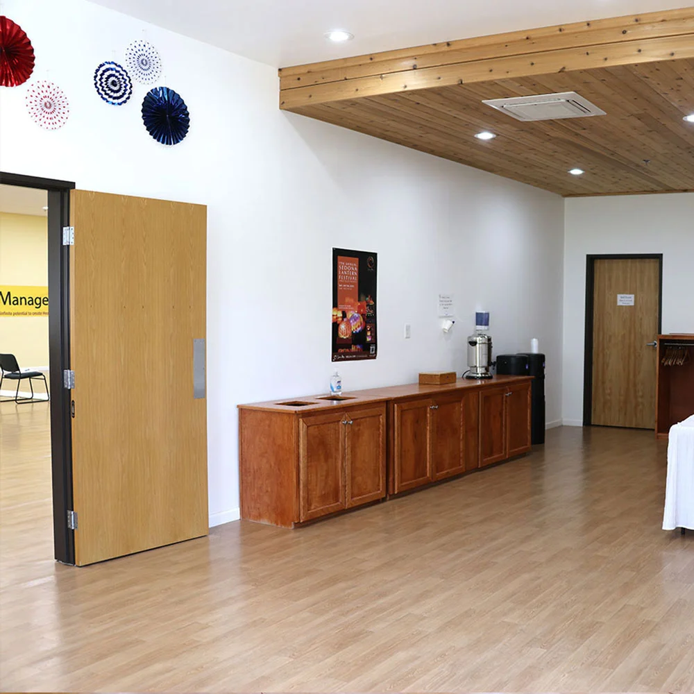 A serene, modern space with wood floors, white walls, wooden ceiling, cabinets, water dispenser, art accents, and open doorway to chairs.