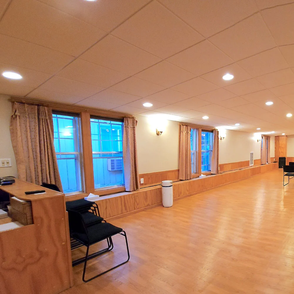 A serene, sunlit space with wood floors, beige-curtained windows, stacked black chairs by a counter, and gentle recessed lighting.