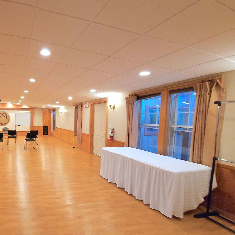A bright, serene room with wooden floors, beige walls, veiled windows, white-clothed table, scattered chairs, and a clothing rack.