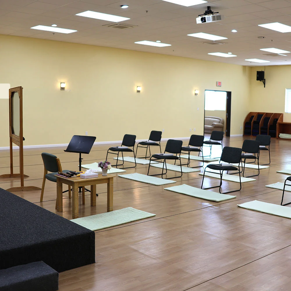 Sunlit room, chairs and mats in tidy rows face a small stage with table, music stand, lectern—inviting peaceful gathering.