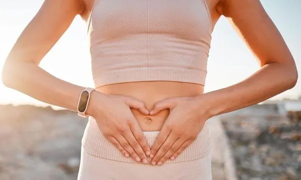 A woman in a beige sports outfit stands outdoors, hands over her belly in a heart shape. Sunlight glows; her wrist shows a fitness tracker.