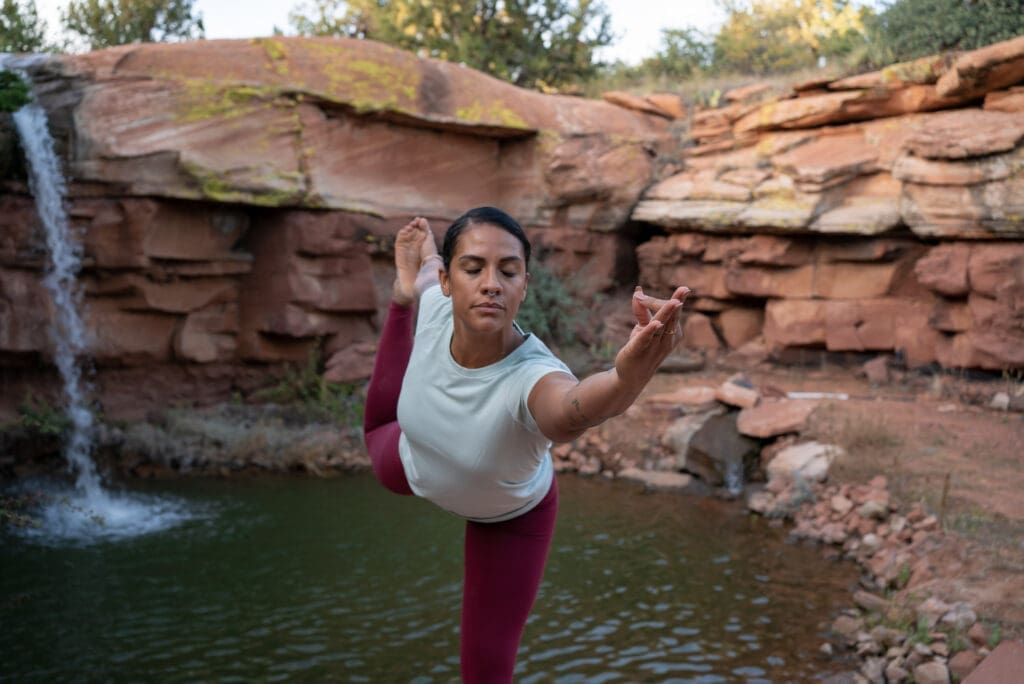 197-DSC02140 A woman balances in yoga by a tranquil pond, red rock cliffs and a waterfall behind her, serene in white shirt and red leggings.