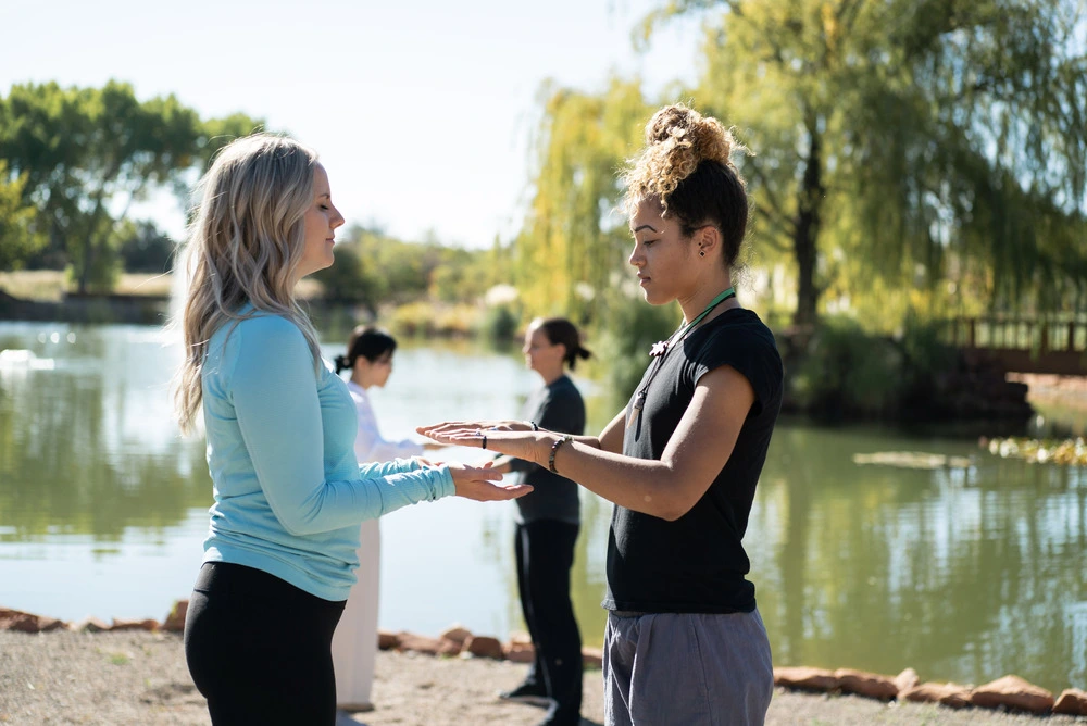 FTS_FAQ By a tranquil lake, two women in meditative stillness face each other, hands extended; others gently flow behind them.