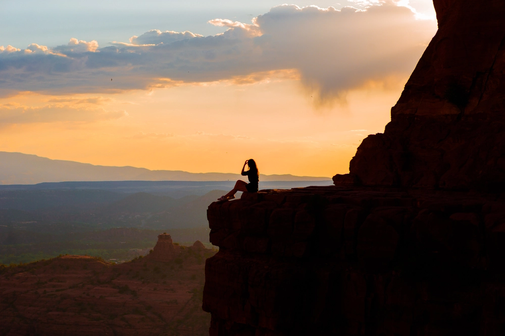 FTS_What happens During the retr On a rocky cliff at sunset, a lone figure sits in quiet silhouette, framed by an orange sky, distant mountains, and drifting clouds.