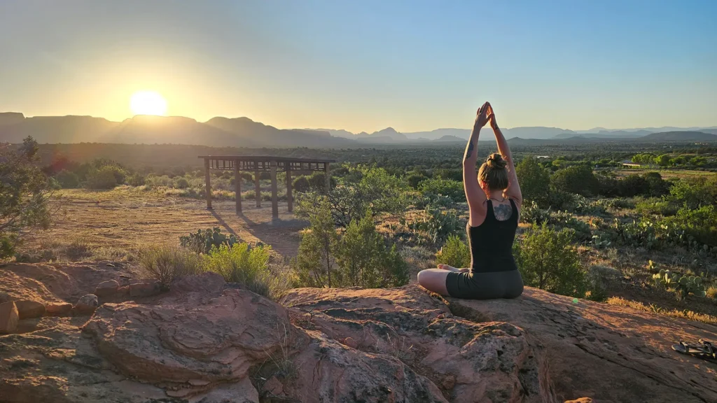 At sunrise, a figure in yoga pose sits on a rocky ledge, gazing over serene desert, mountains, green shrubs, and long-shadowed wood.