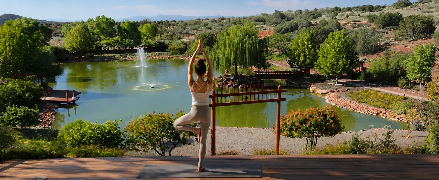 Yoga Retreats in Sedona A person in tree pose practices yoga on a deck by a peaceful pond and fountain, embraced by lush green trees under a calm blue sky.