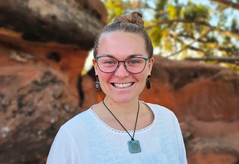 Yoga-Trainer-1000-686 A smiling person with glasses and light brown hair in a bun stands outdoors, embraced by red rocks and gentle greenery behind them.