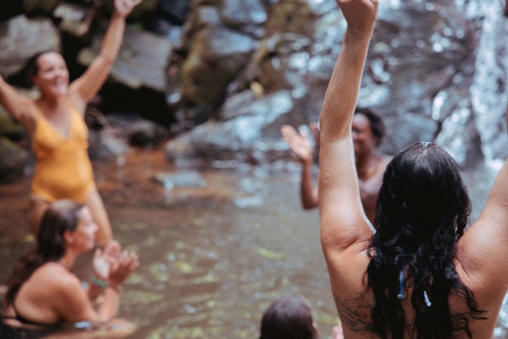 People gather in a serene natural pool by rocks, raising arms, clapping, smiling—immersed in joyful outdoor harmony together.