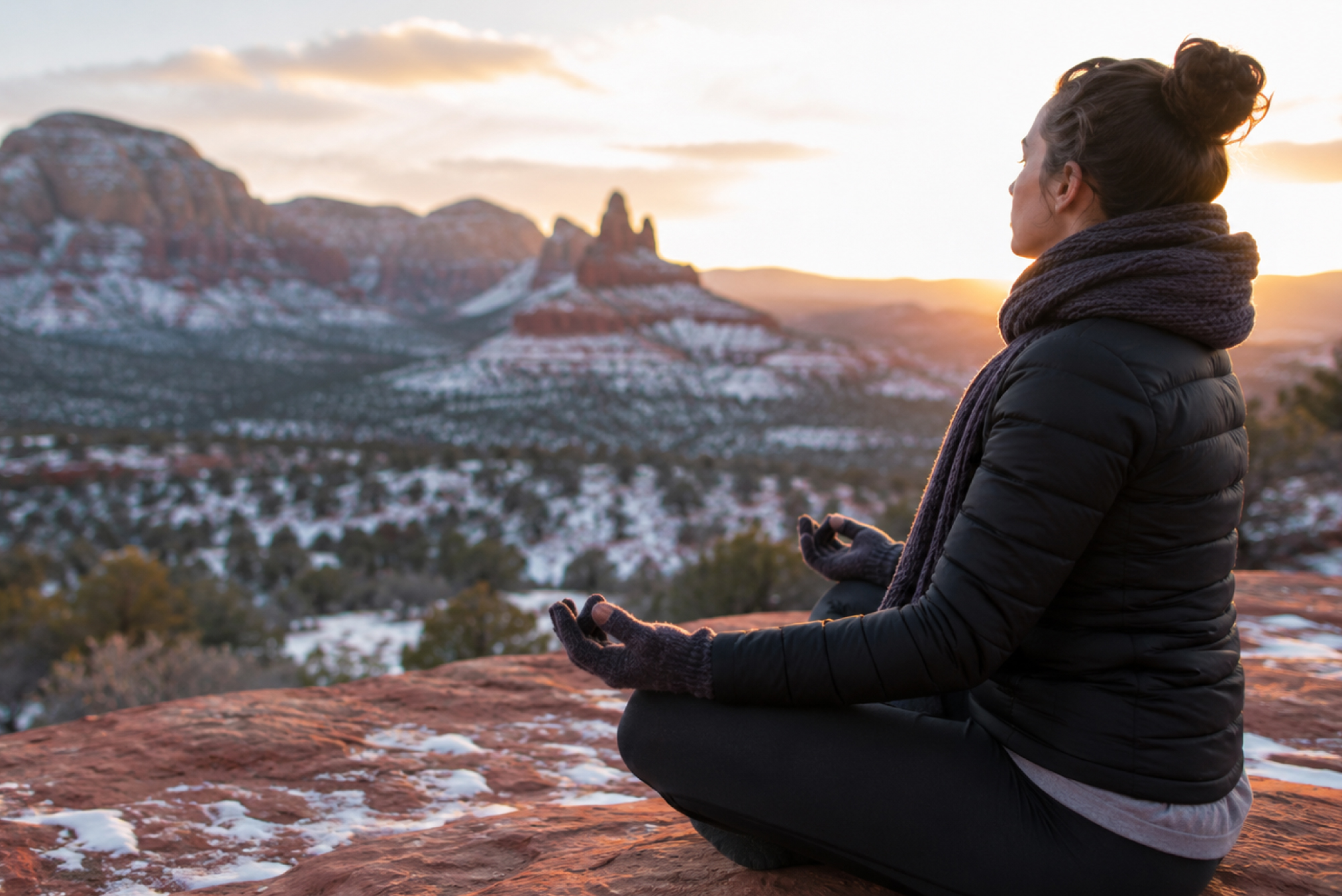 SM-Program-Images-NY-1 A person sits cross-legged in meditation on a rocky ledge, gazing over a tranquil valley where red rock formations rise amid patches of gentle snow, all illuminated by the golden light of sunrise or sunset.