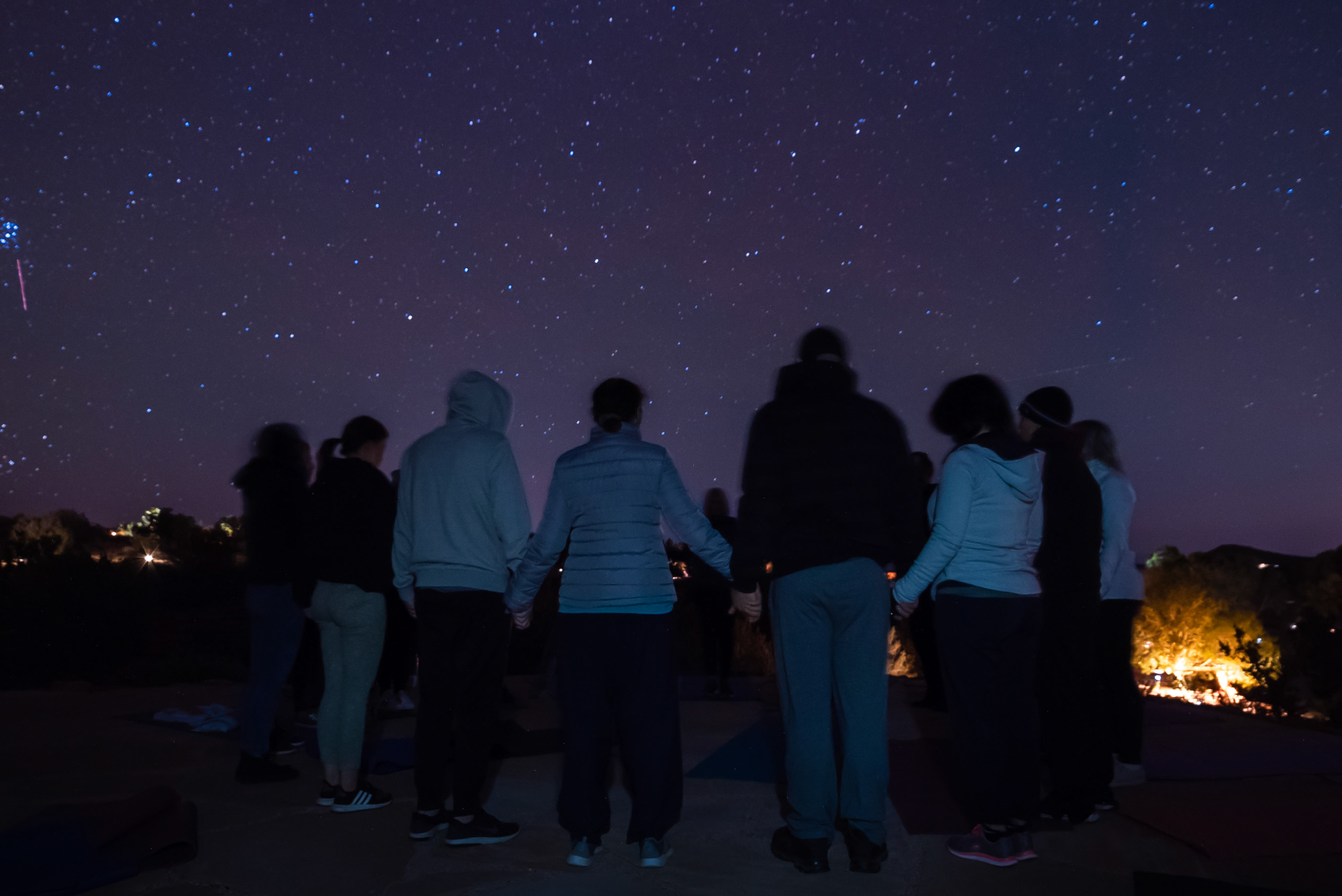 SM-Program-Images-NY-2 Under a vast starry night sky, a group of people join hands, forming a circle. Their silhouettes are gently illuminated by the soft glow of distant lights, framed by the quiet presence of trees in the background—a moment of unity and peace beneath the universe's embrace.