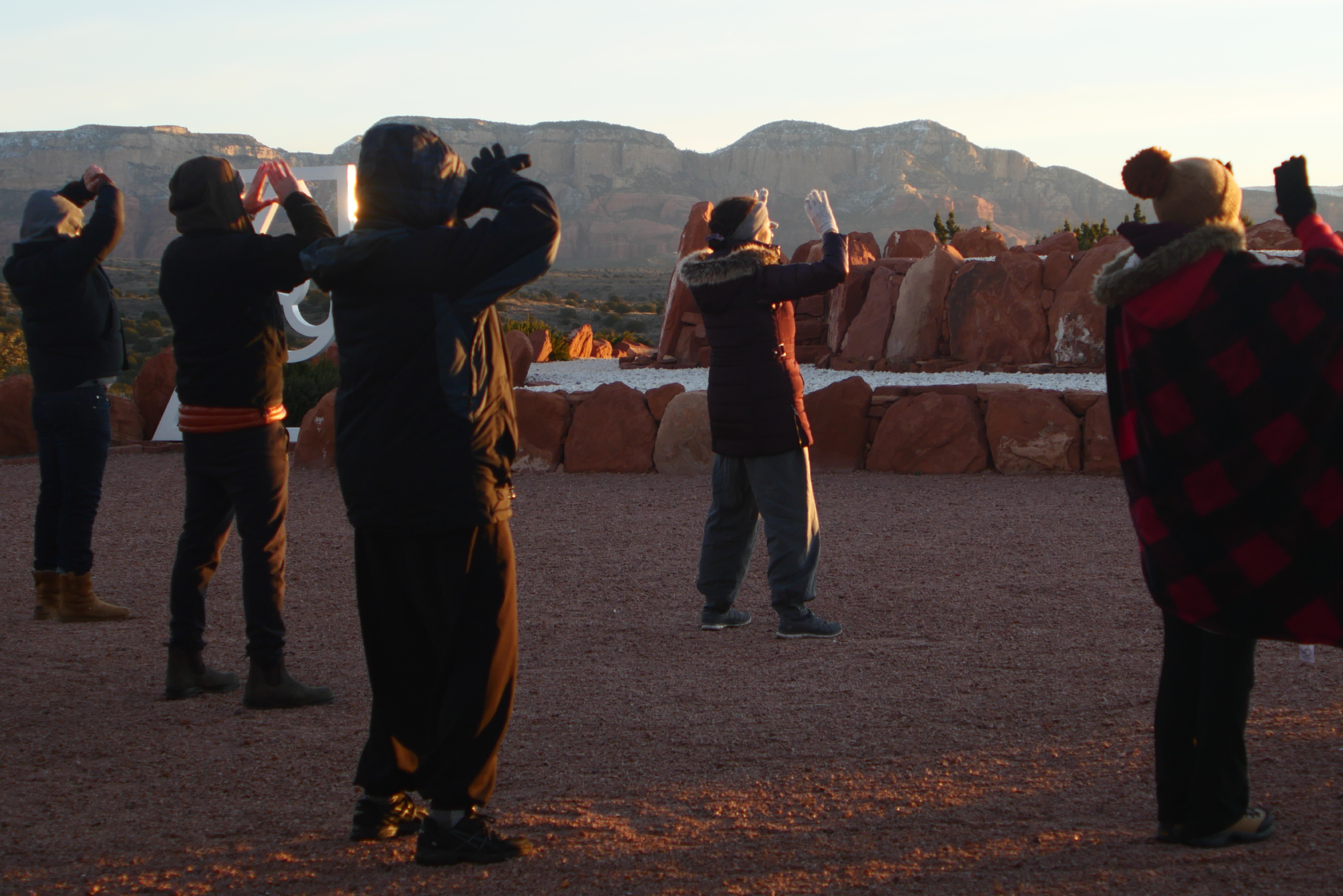 SM-Program-Images-NY-3 Six people gather on gravel beneath the vast sky, warmly bundled against the chill as they lift their arms upward. Behind them, Sedona’s signature red rocks and distant mountains glow gently in the golden light of dawn or dusk.