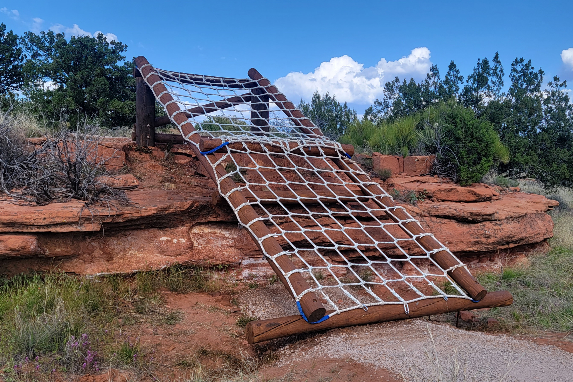 SM-Program-Images-v10 Amid rugged red sandstone, a rope net climb made from sturdy logs and white rope stands, surrounded by desert shrubs and watchful trees beneath a spacious blue sky adorned with gentle clouds.