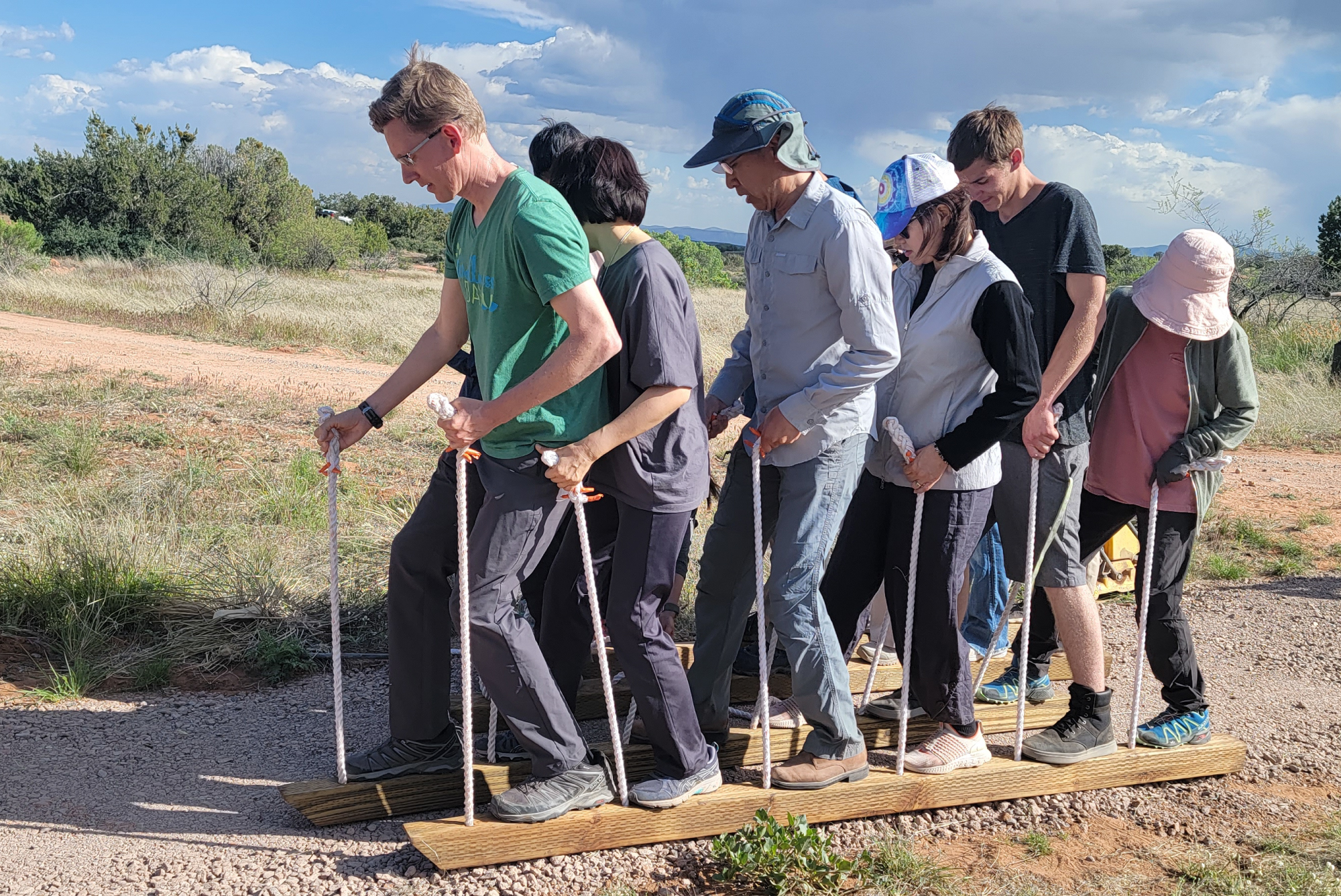 SM-Program-Images-v11 Eight people stand together on two long wooden boards, each holding ropes, moving in unison as they walk across a gravel path outdoors. Around them stretch open grassy fields and the gentle presence of trees, all beneath a partly cloudy sky that breathes possibility into the scene.