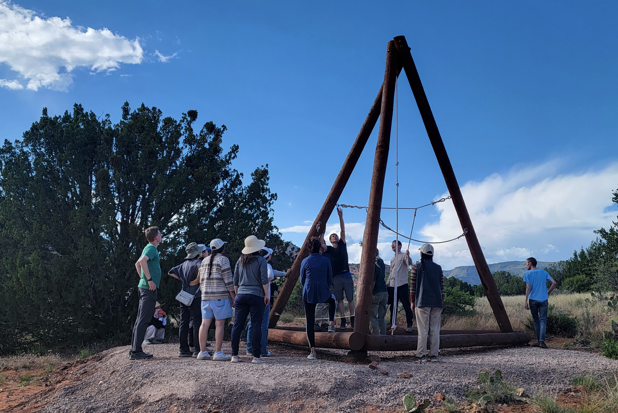 SM-Program-Images-v12 A gathering of people encircles a large outdoor pendulum structure resting on a gravel clearing, where trees whisper nearby and distant mountains rise beneath a sky softly veiled with drifting clouds.