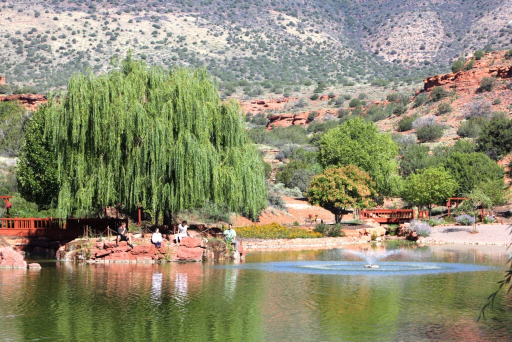 SM-Program-Images-v7 A tranquil pond with a shimmering fountain rests amid vibrant green trees and majestic red rock formations. People pause along the shore, soaking in the serene beauty. In the background, a wooden bridge and walkway invite gentle exploration beneath radiant sunlight.