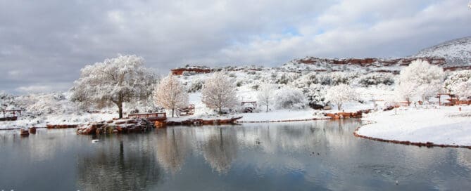 A tranquil snowy landscape stretches before you, where gentle trees and rolling hills mirror themselves in the serene stillness of a calm lake. Above, clouds drift through a softly lit sky, and nearby along the shore, simple benches and wooden structures invite quiet contemplation amidst nature’s winter embrace.
