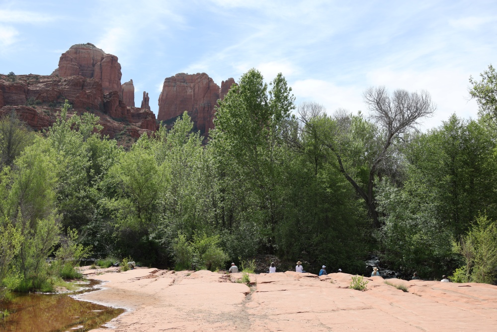 vortex hike Majestic red rocks ascend behind lush, vibrant trees, grounding the landscape in serene energy. In the foreground, several people sit peacefully on flat stones warmed by sunlight, while a gentle stream flows quietly to the left beneath a sky adorned with soft clouds.