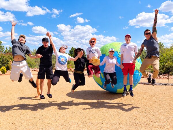 A group of people in casual clothing leap for joy before a large globe, embodying the spirit of Sedona Mago.